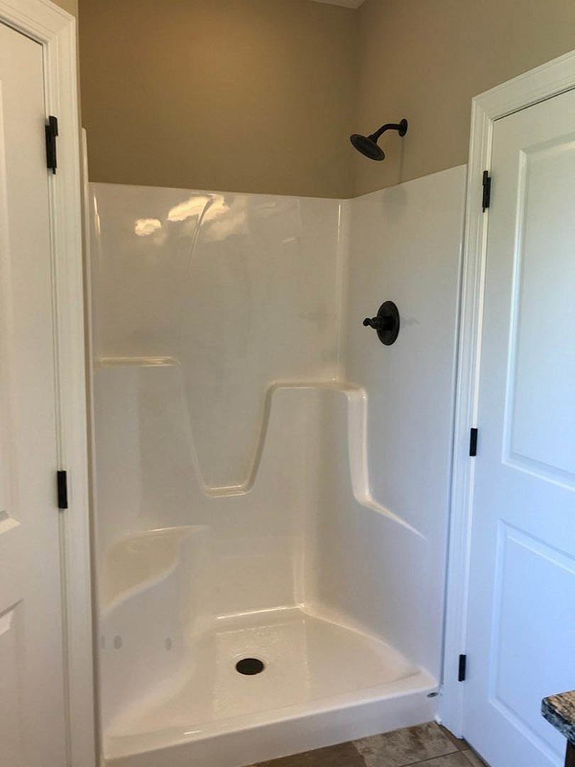 White tiled shower with black metal shower head and handle, built-in drain, and adjacent white door featuring a black handle