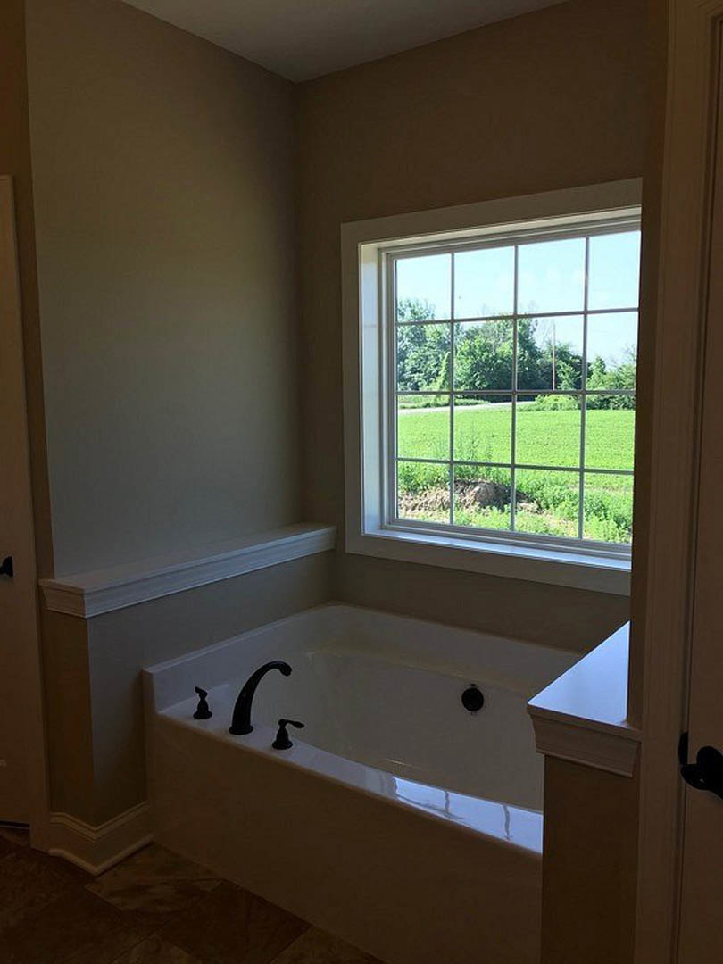 Freestanding bathtub beneath large window overlooking grassy field, white tile walls, chrome faucet, modern bathroom interior
