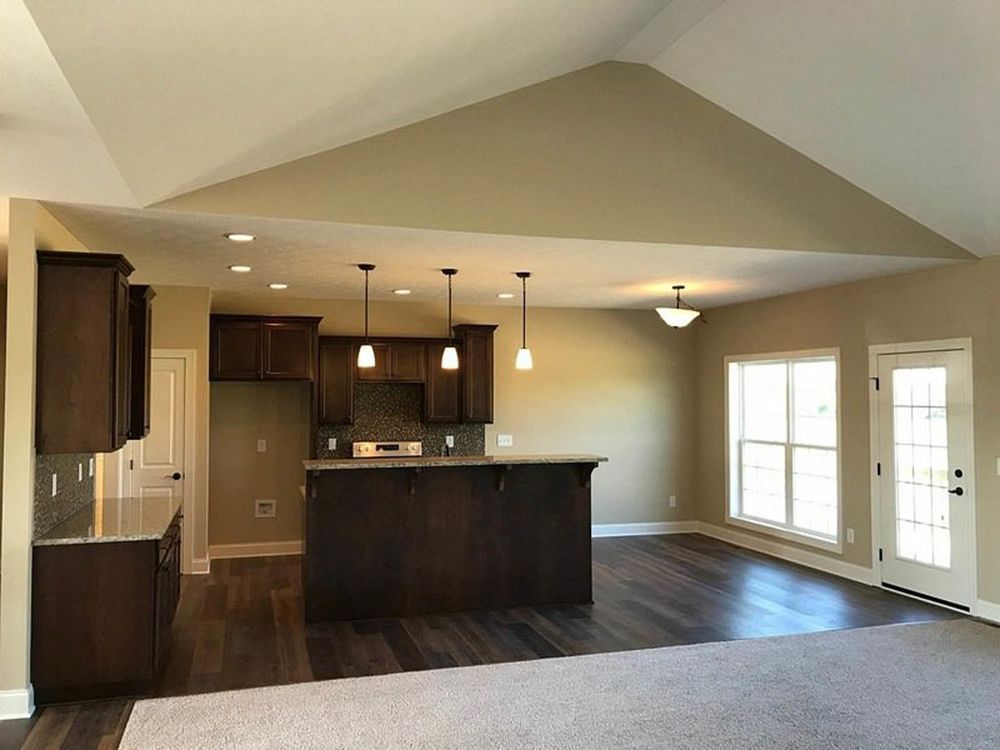 Open-concept kitchen and living room with high ceiling, dark brown countertop, white door with glass panels, carpeted flooring, large window, and modern light fixture