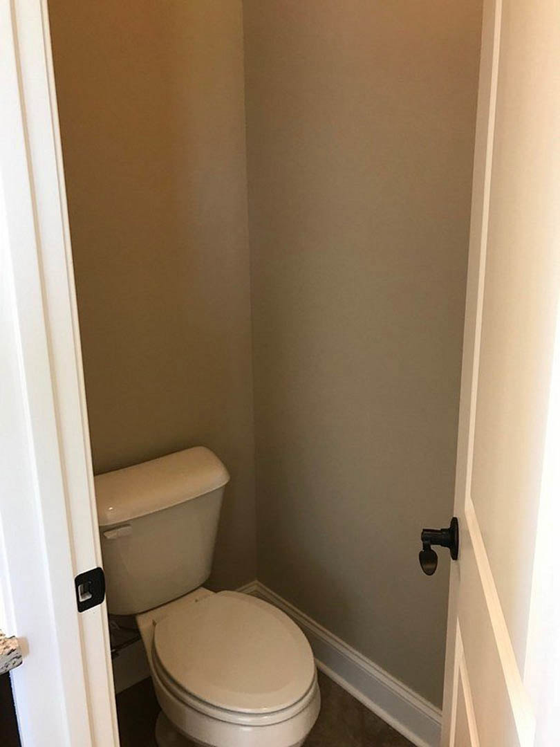 White toilet with lid up against tiled wall in compact bathroom, chrome flush handle on tank, light-colored floor tiles, minimal decor.