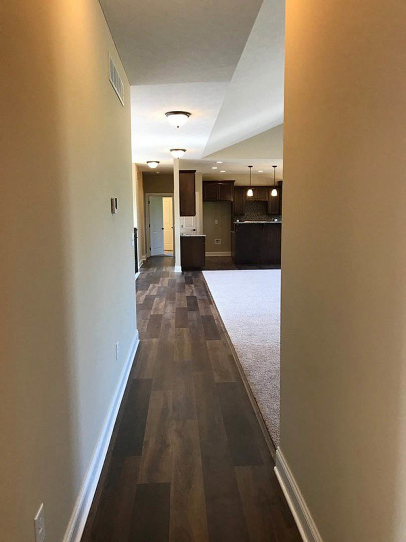 Hallway with light hardwood flooring, white walls, white door with black knobs, black rectangular wall fixture with white border, and close-up views of carpet and white surface