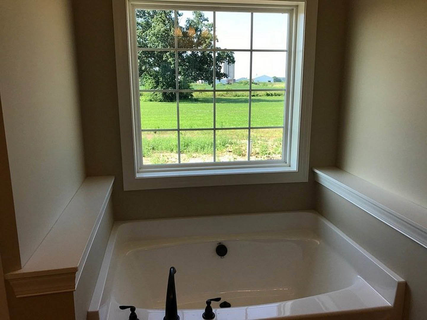 Freestanding white bathtub beneath large window, chrome faucet, view of grassy field and trees outside, light-toned walls, minimalist bathroom design