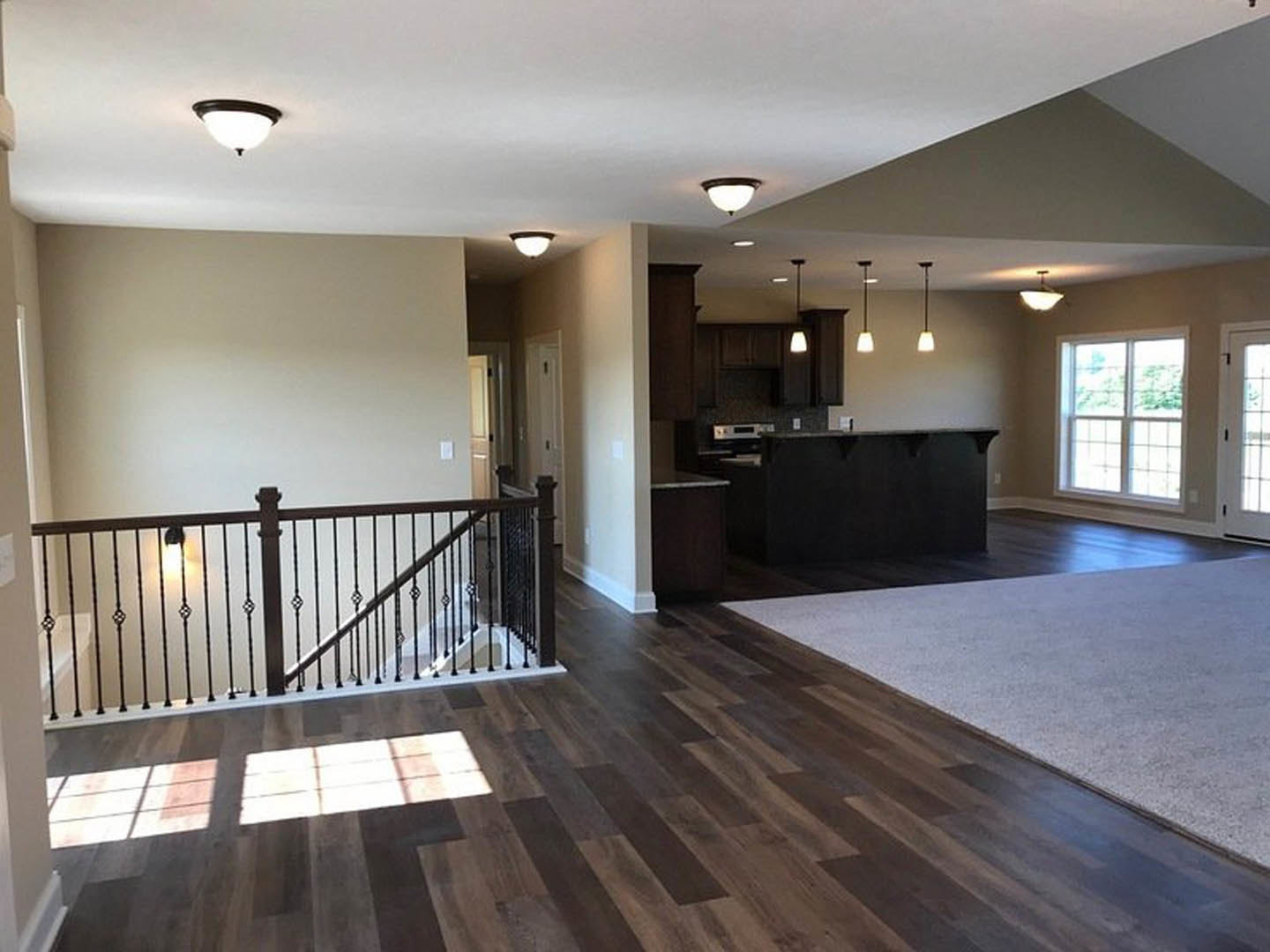 Open-concept living room with wood flooring, carpeted area, white-framed window, staircase featuring wood steps and railing, and adjacent kitchen with black and white countertops