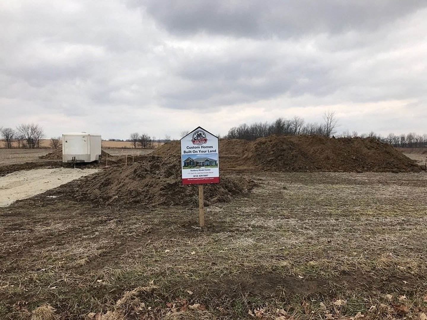 Wooden real estate sign with house logo stands in grassy field, white trailer and distant tree visible under partly cloudy sky.