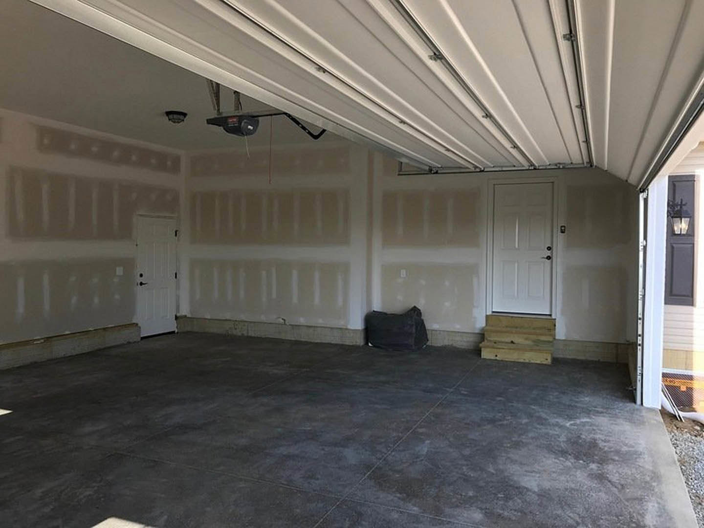 Garage with smooth white walls, polished concrete floor, white door featuring black knobs, black pillow resting on a couch, close-up of wooden step, grey tile floor with black bag