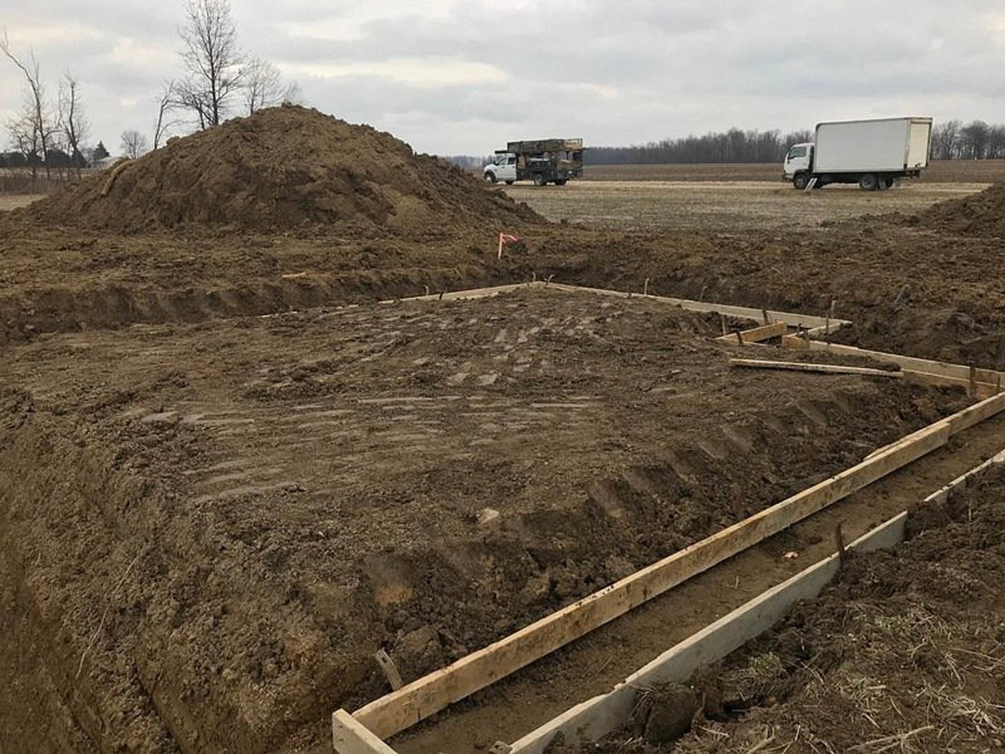Dirt piles and wood planks on a residential lot, white truck parked on a dirt road, cloudy sky and scattered trees in the background