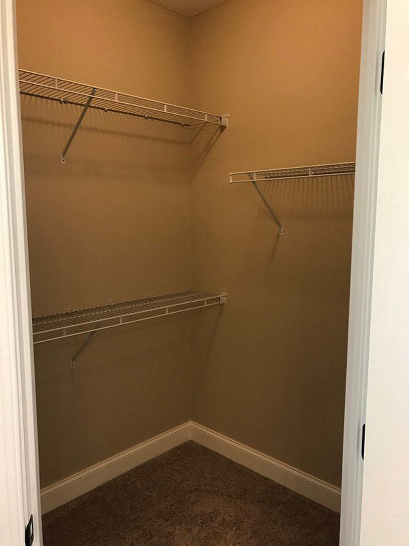 Walk-in closet with white walls, built-in metal shelves and hanging rail, brown carpet flooring, white door frame visible in the corner.