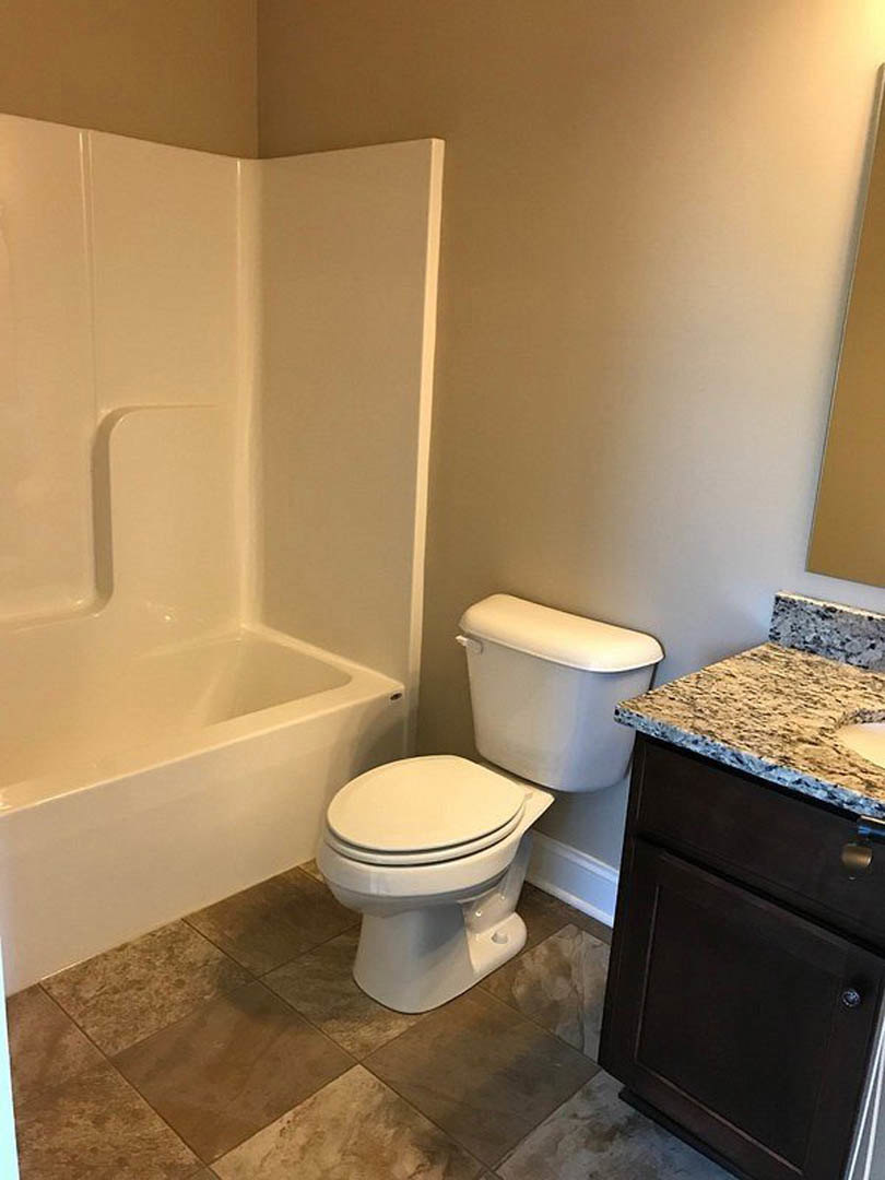 White toilet beside a built-in bathtub, light tile flooring, wood vanity with stone countertop, neutral wall tiles, chrome fixtures