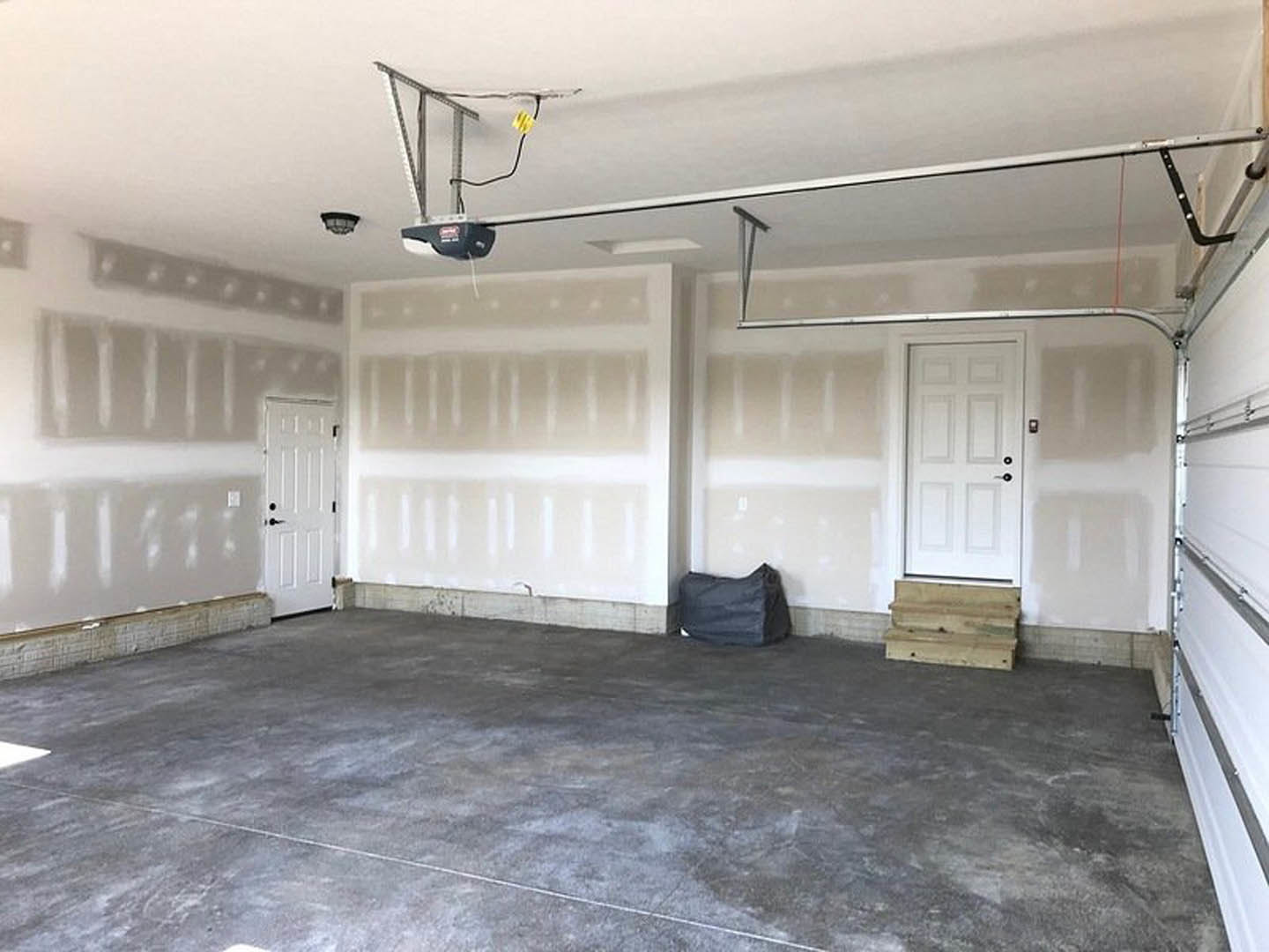 White walls and concrete floor in an empty room, black bag on a table, white door with black knobs, wood plank resting on window sill, bag placed on the ground.