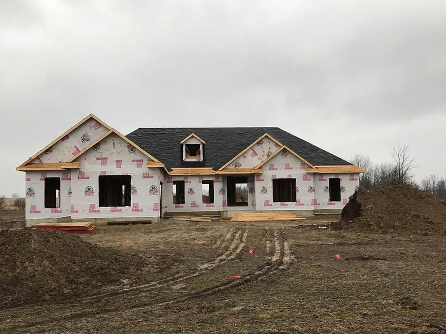Modern house under construction featuring black roof, exposed window and doorway openings, surrounded by dirt and unfinished ground under cloudy sky