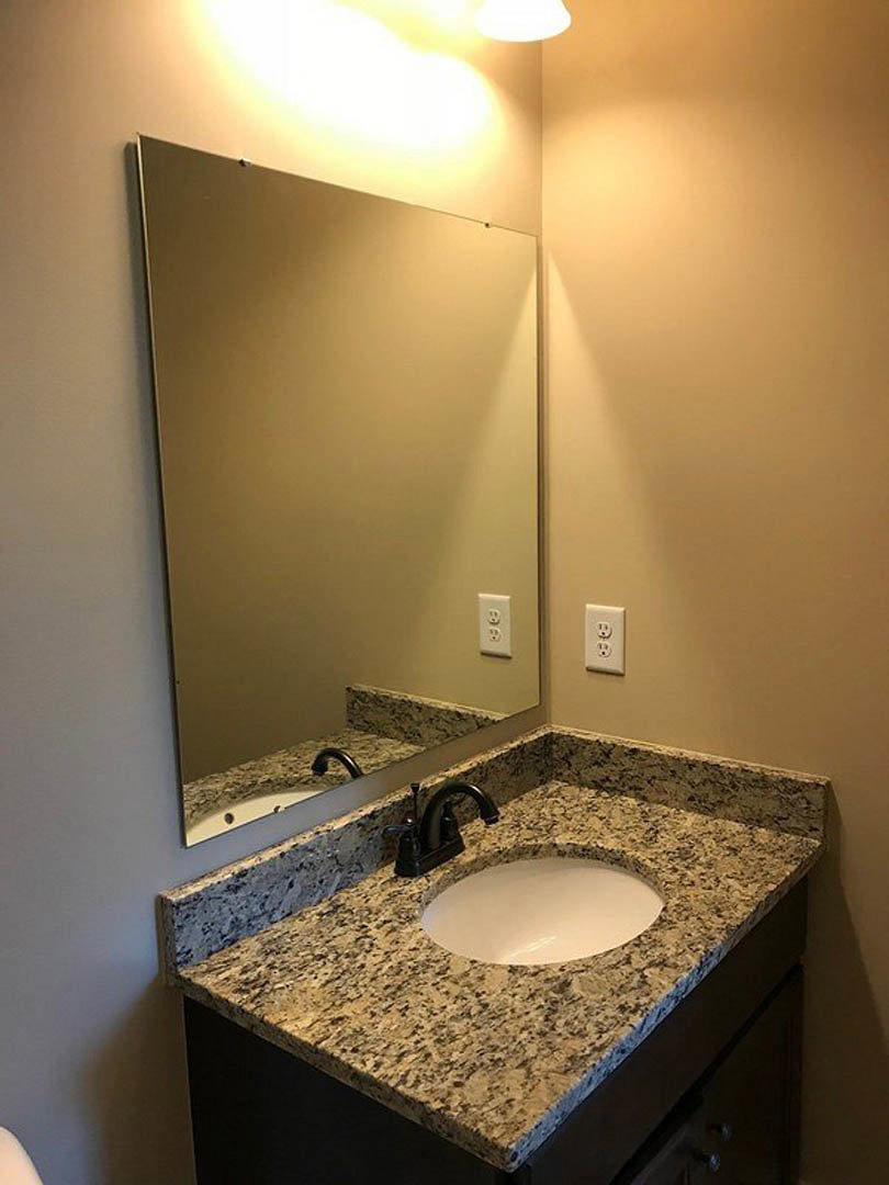 White round sink with black faucet beneath large rectangular mirror, wall-mounted light fixture above, gray tile backsplash, bathroom cabinet below.