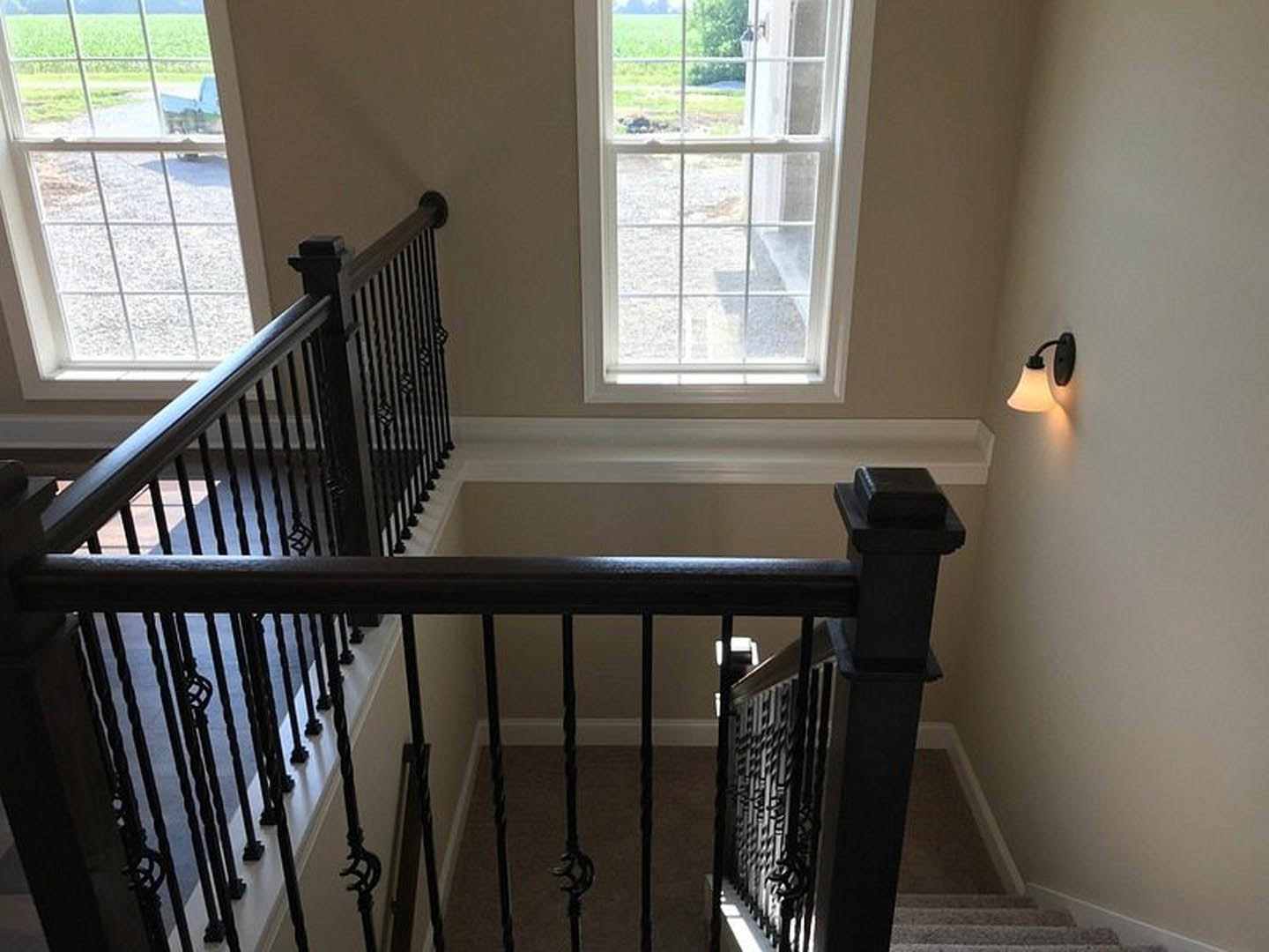 Staircase with black iron railing, large windows overlooking grass and gravel path, wall-mounted light fixture, white walls, hardwood flooring