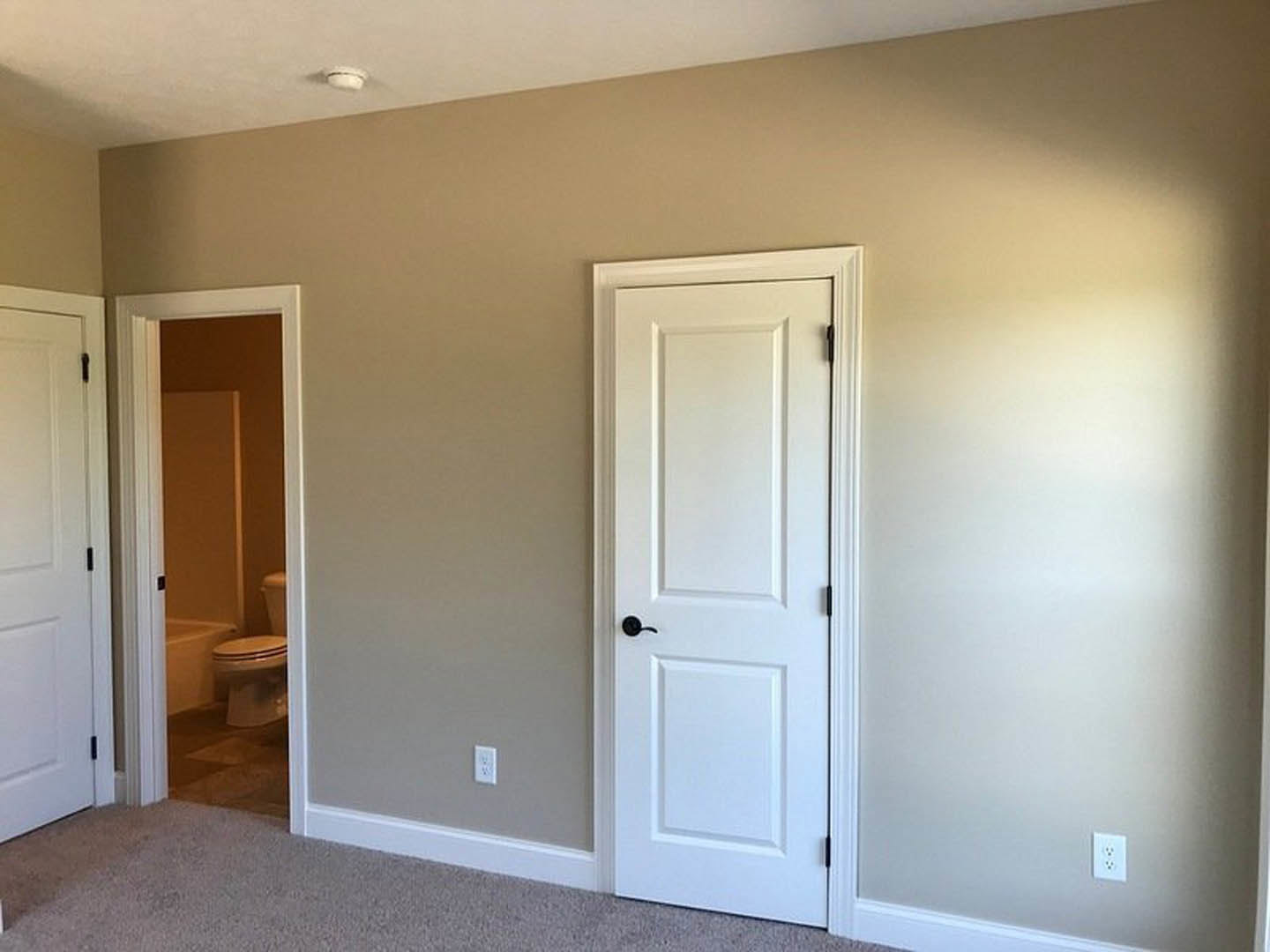 White paneled door with black handle next to a white outlet, opening into a bathroom with a closed toilet seat and bathtub, set against light walls and flooring.