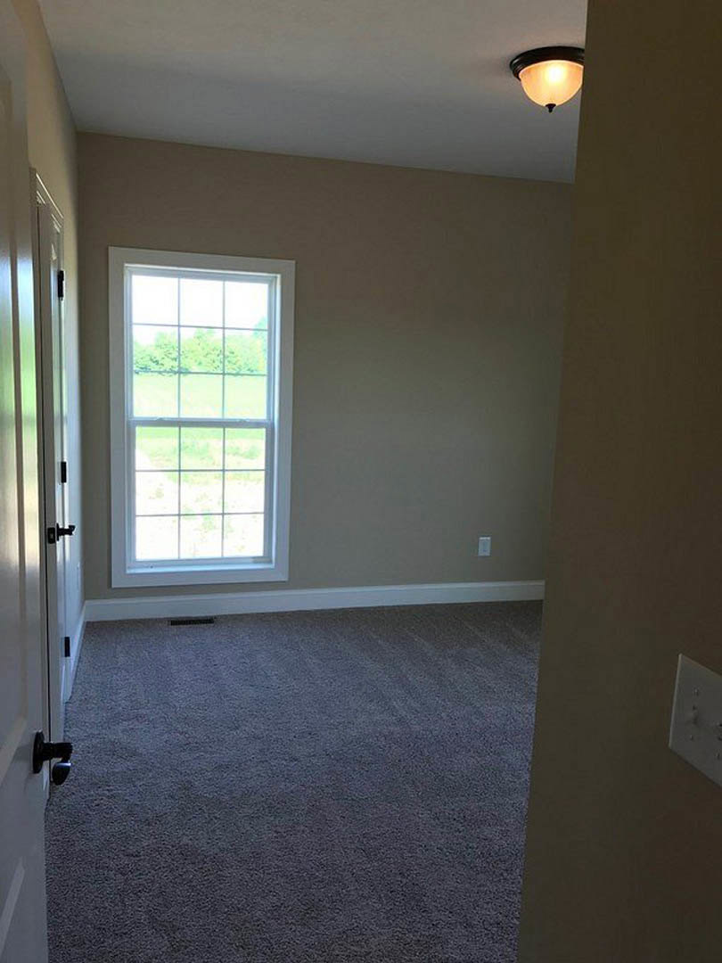 Carpeted bedroom with large window overlooking grassy field, white walls, ceiling light fixture, and wooden door