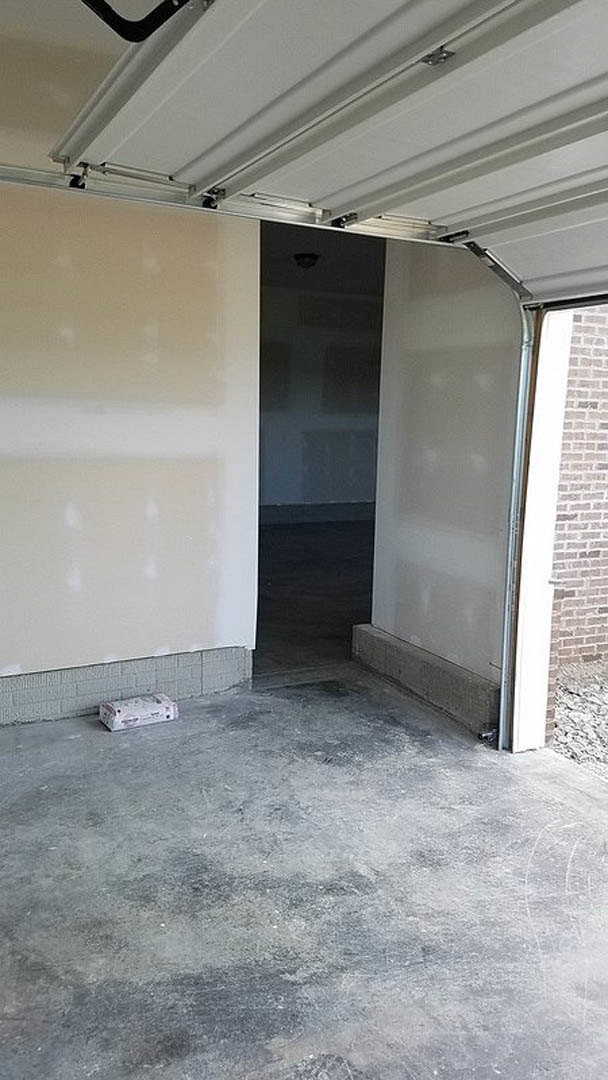 White garage door set in a plastered concrete wall, small utility box mounted to the right side, smooth indoor flooring and ceiling visible.