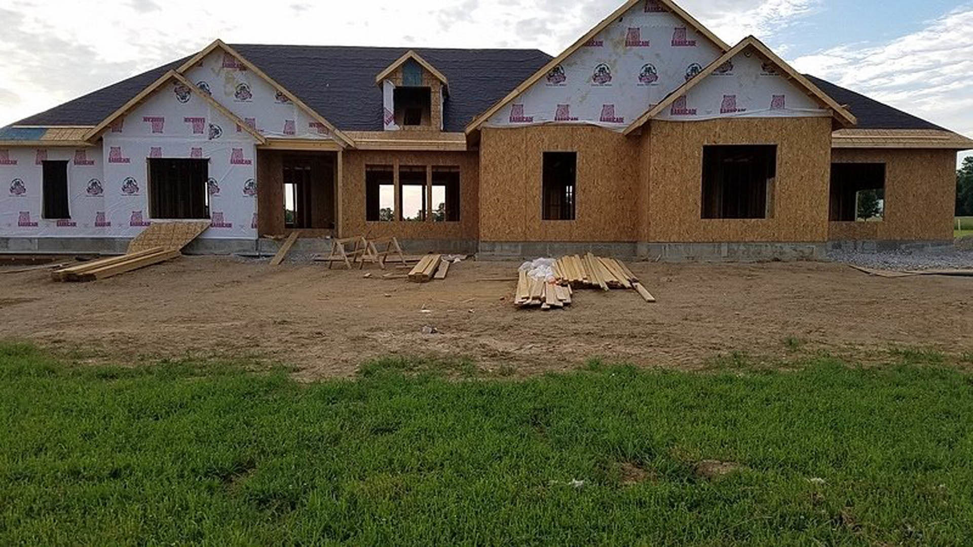Partially built house with blue roof, dark-framed windows, pile of wood on grassy yard, cloudy sky overhead