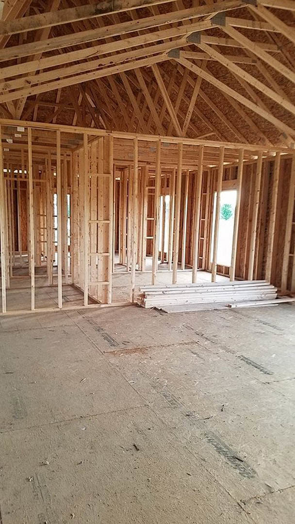 Exposed wood framing with visible beams, concrete floor, unfinished staircase, window opening, roof structure, and tire tracks in sandy ground