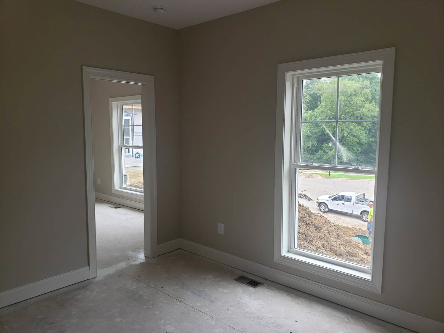 Bright room with plaster walls, two large windows with white frames, view of dirt road outside, white truck parked, hose spraying water on ground, man visible in background.