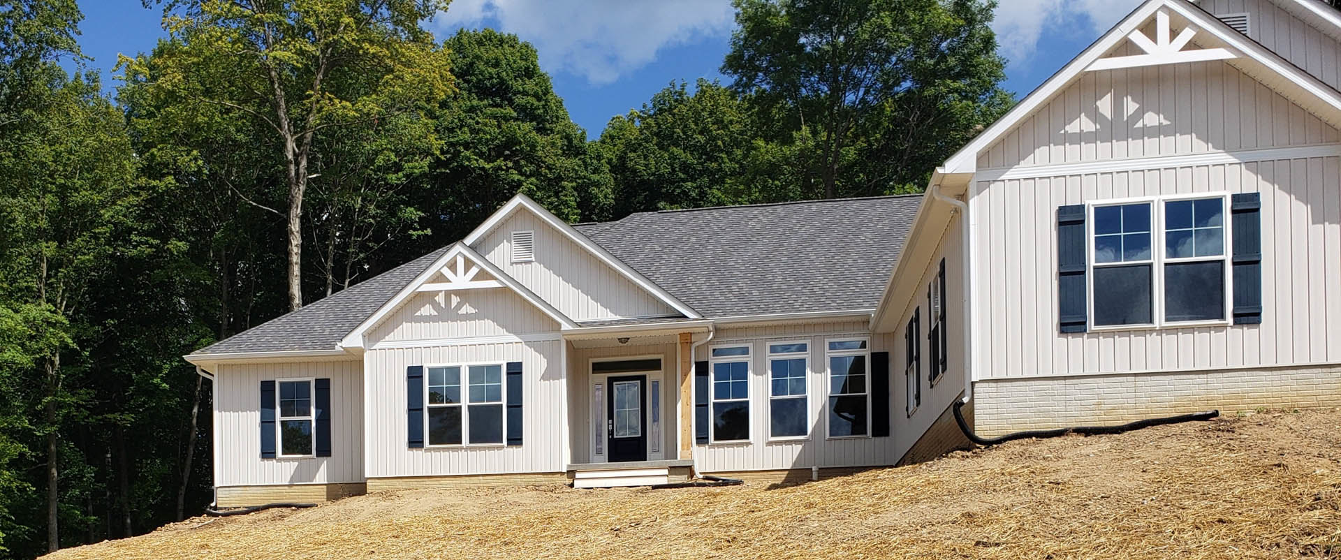 Modern cottage-style home with white siding, black front door, large window with white frame, set against a grassy hill and mature trees
