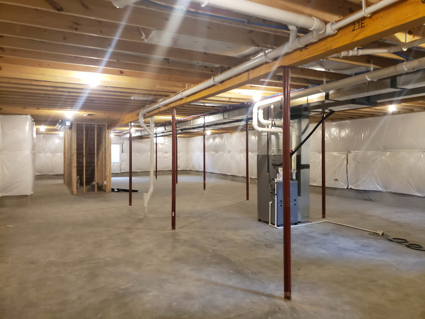 Unfinished basement with exposed steel beams, concrete floor, visible plumbing pipes, white drywall, and overhead lighting