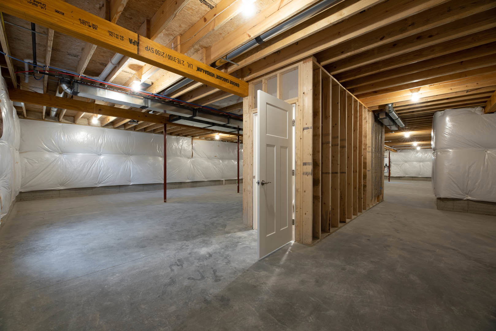 White door open in unfinished basement room with exposed wood ceiling beams, visible pipe, and building insulation; white mattress rests on floor.