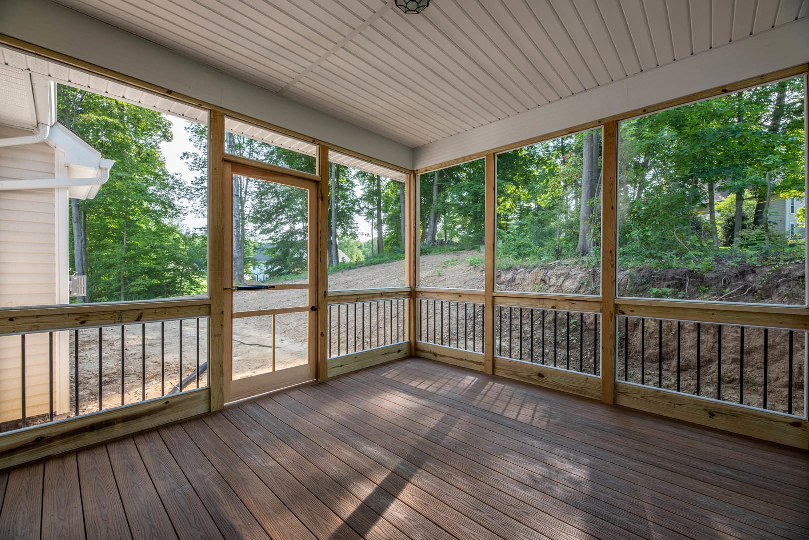 Wooden porch with white ceiling, light fixture, and railings overlooking trees and distant hill; window frames view of neighboring house.