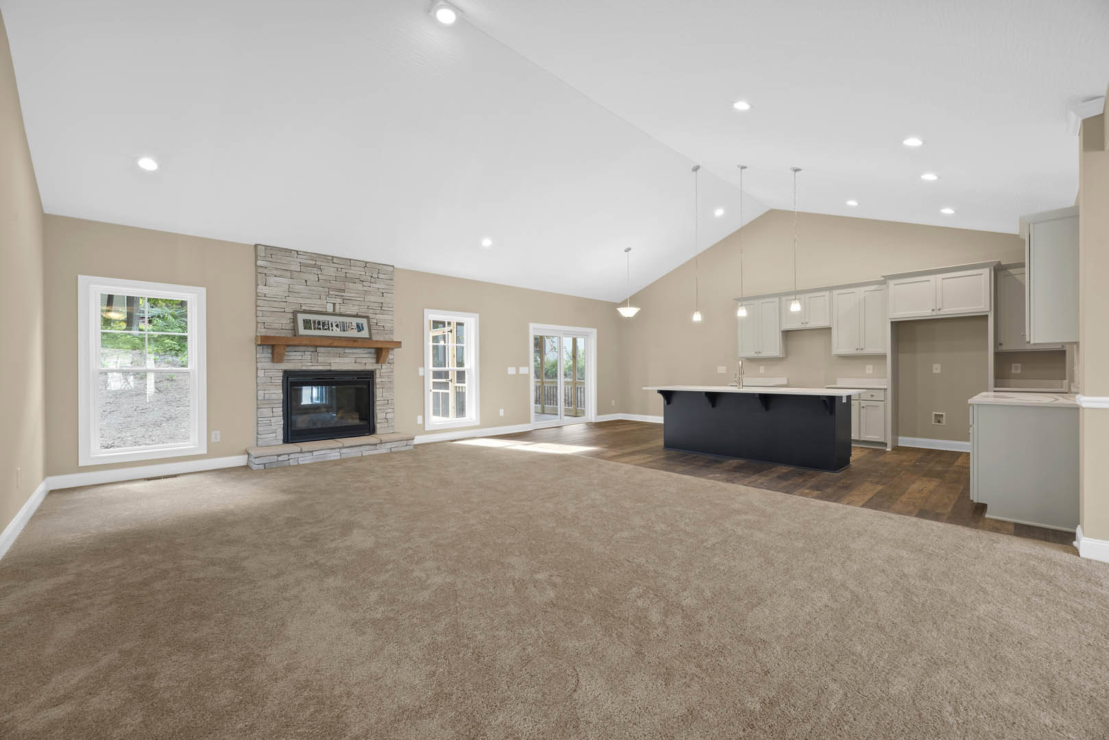 Spacious living room with carpeted floor, central fireplace, adjacent kitchen featuring black cabinetry with white countertops, large windows overlooking yard, and white door with