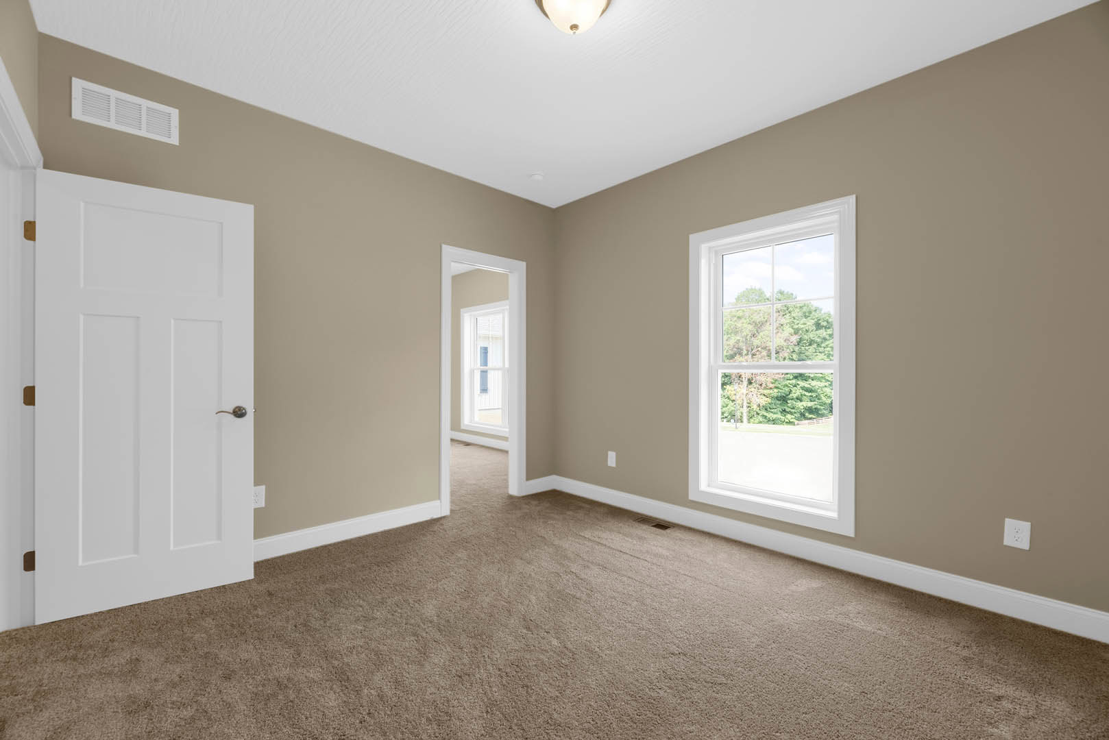 Beige-walled room with white trim, carpet flooring, white door featuring a silver handle, window showing trees outside, wall vent, and white electrical outlet