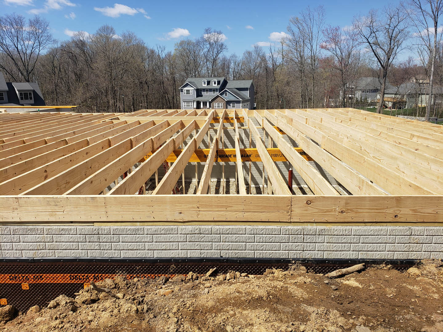 Wooden framing of a house under construction on a dirt lot, with a finished home and trees visible in the background.