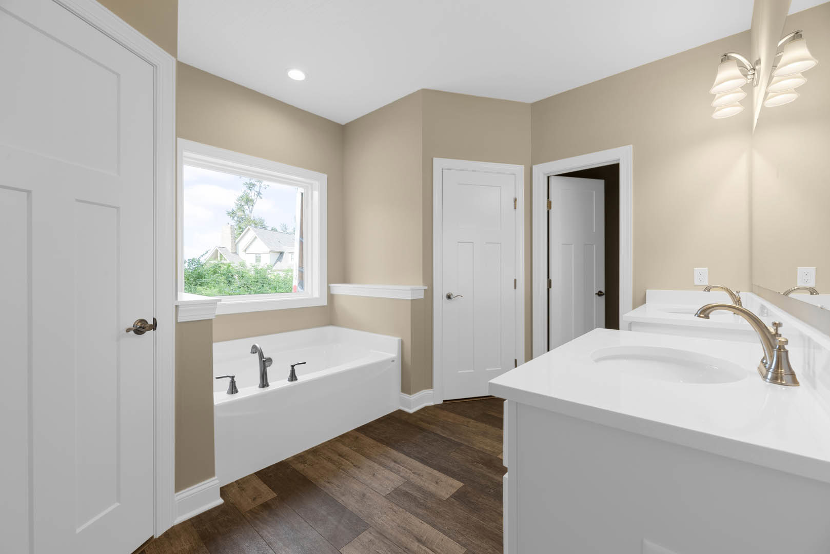 Bathroom with dual white sinks, freestanding white tub, wood flooring, silver faucet fixtures, white door with silver handle, window overlooking neighboring house, modern light