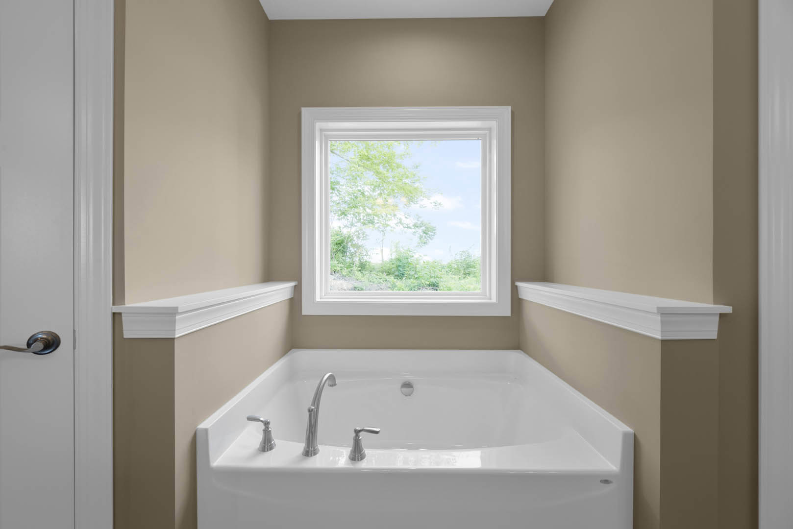 Freestanding white bathtub with chrome faucet set against tiled bathroom wall, large window overlooking trees, white molding detail, and towel hook visible nearby