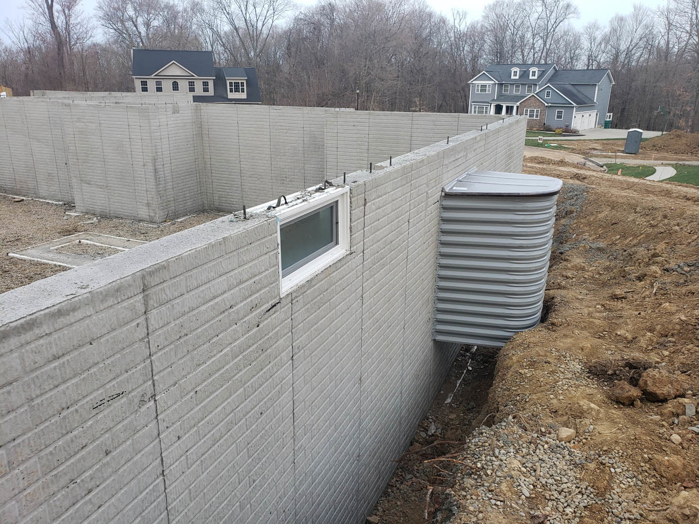 Concrete exterior wall with two rectangular windows, surrounded by dirt ground and sparse vegetation, metal barrel and structure visible nearby.