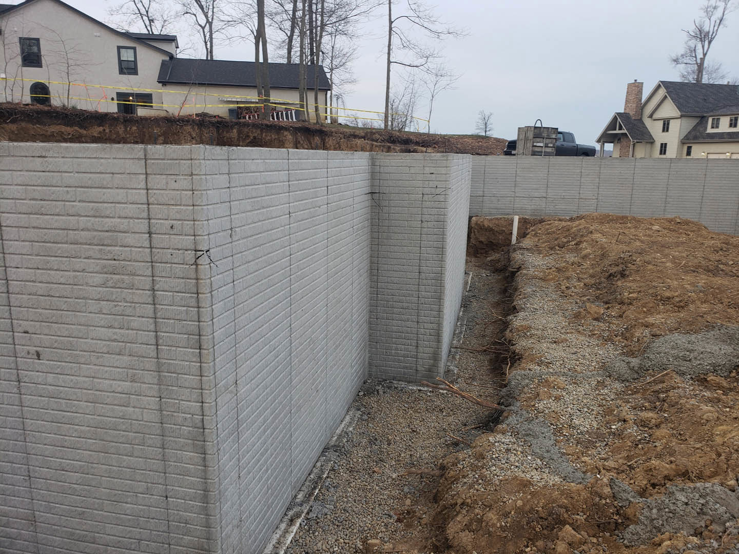 Concrete exterior wall with visible corner crack, yellow caution tape stretched across dirt and gravel construction site, chimney and fence in background, trees and sky visible