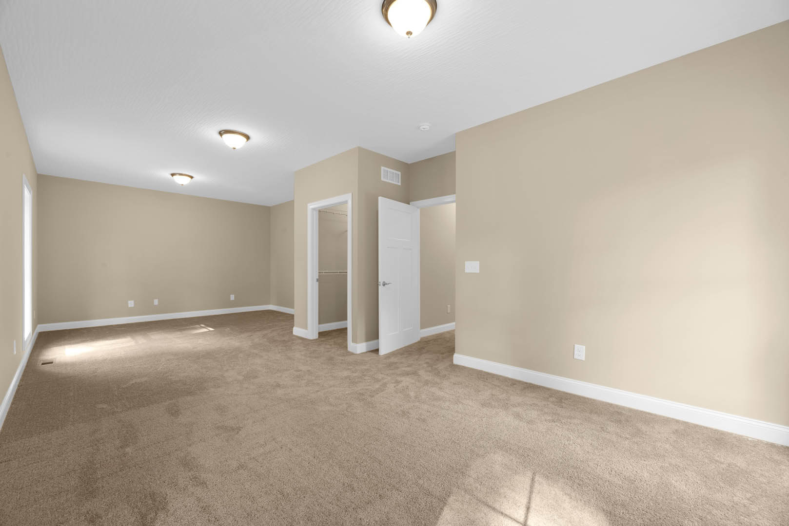 Beige-walled room with white door featuring silver handles, open to reveal bathroom; ceiling-mounted light fixture and carpeted flooring visible