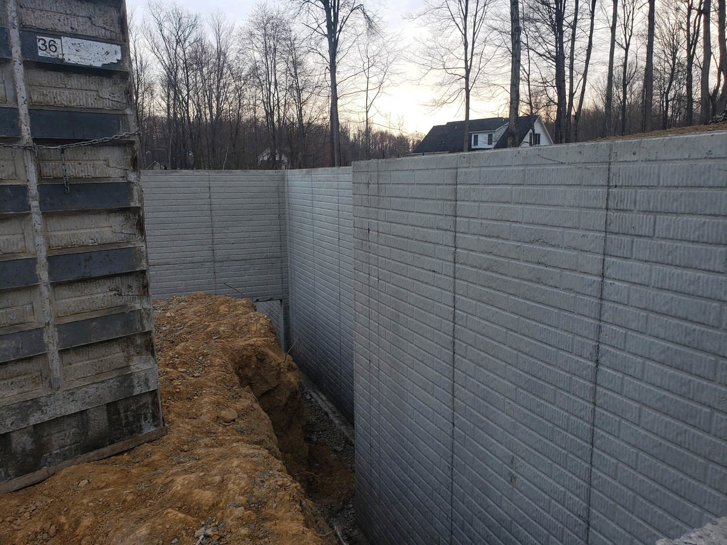 Grey concrete wall with a circular hole in the ground, house with white roof and trees visible in the background