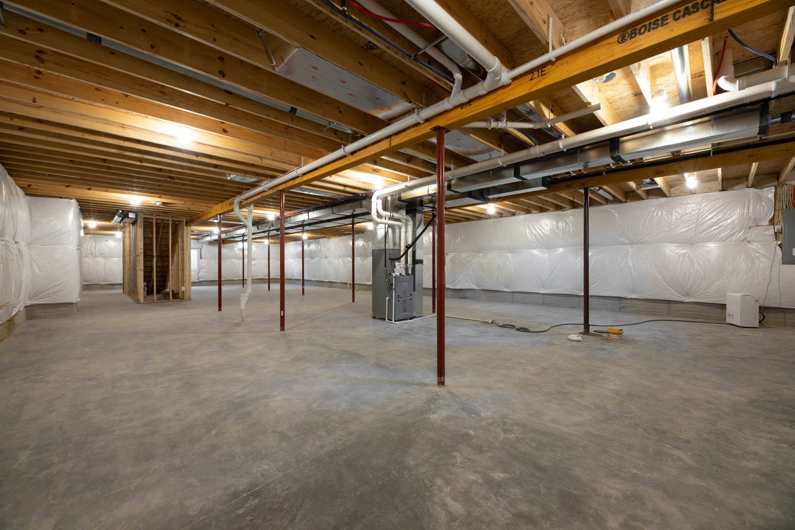 Basement with exposed steel pipes and beams on ceiling, grey concrete floor, red support poles, white bags stacked against wall, close-up of warning sign