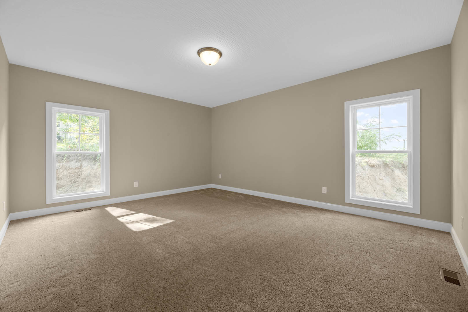 Beige-walled room with white-framed windows, carpeted floor, ceiling light fixture, and sunlight streaming in; window offers view of a dirt hill.