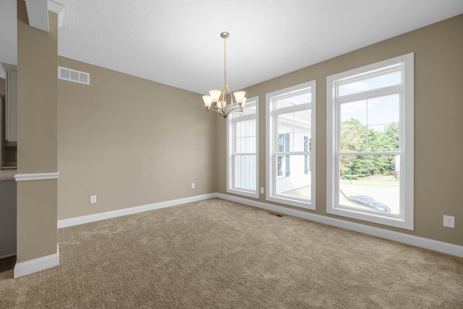 Carpeted living space with large windows framed by white trim, modern chandelier hanging from ceiling, wall outlet and air vent visible, trees seen through windows