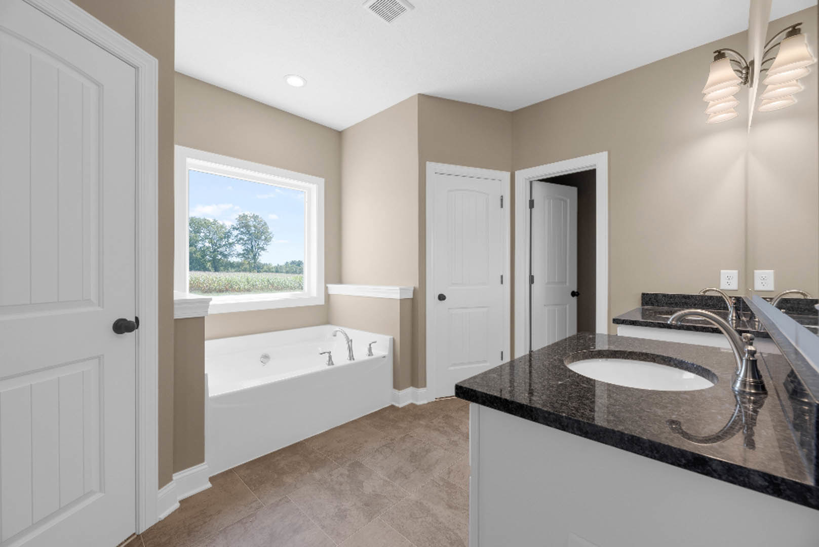Bathroom with freestanding white tub, chrome faucets, brown tile floor, white sink with black hardware, window showing trees, close-up of lamp, white door with black knob.
