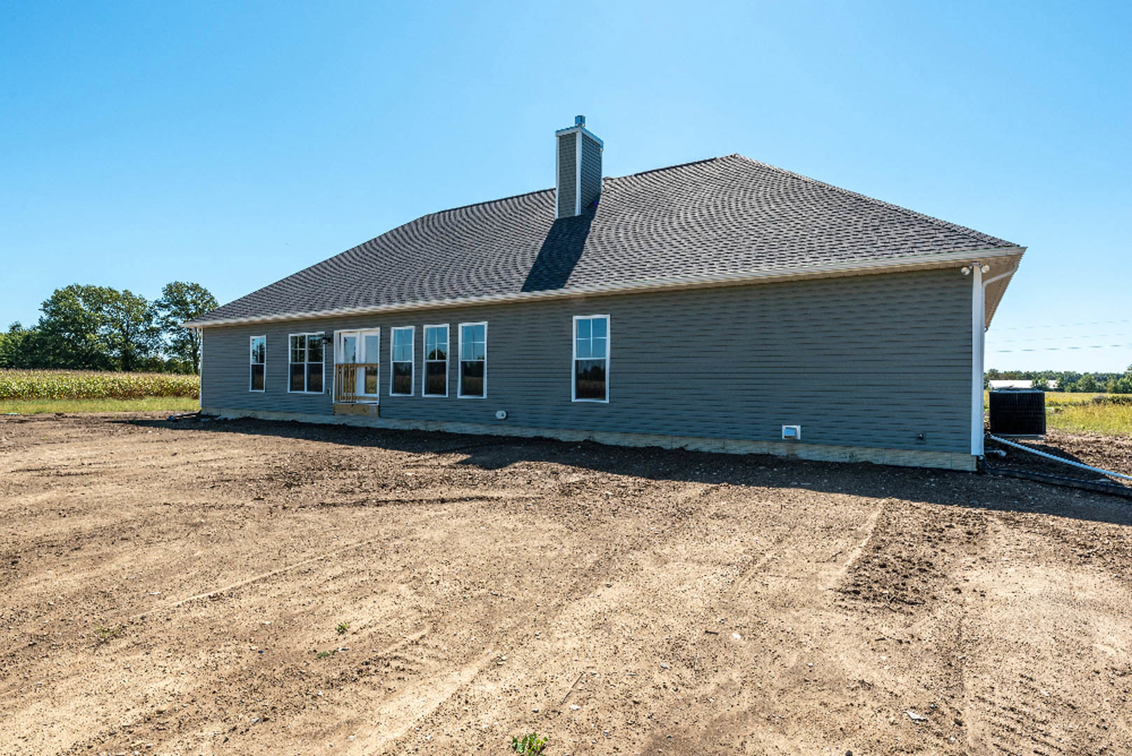 Single-story cottage with blue exterior walls, white-framed windows, chimney on roof, dirt yard in foreground, and trees in sunlight surrounding property.