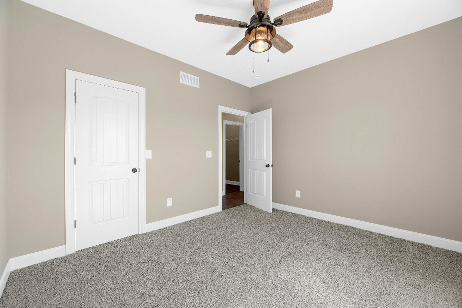 Bedroom with light wood flooring, white plaster walls, ceiling fan with integrated light, white door featuring black handle, and wall-mounted white air vent