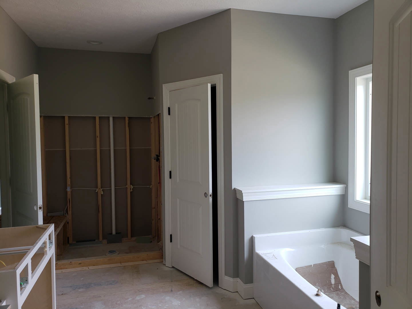 Bathroom with freestanding tub featuring built-in seat, white tile walls, visible plumbing pipes, white door with black handle, metal railing, and white shelving against a plaster