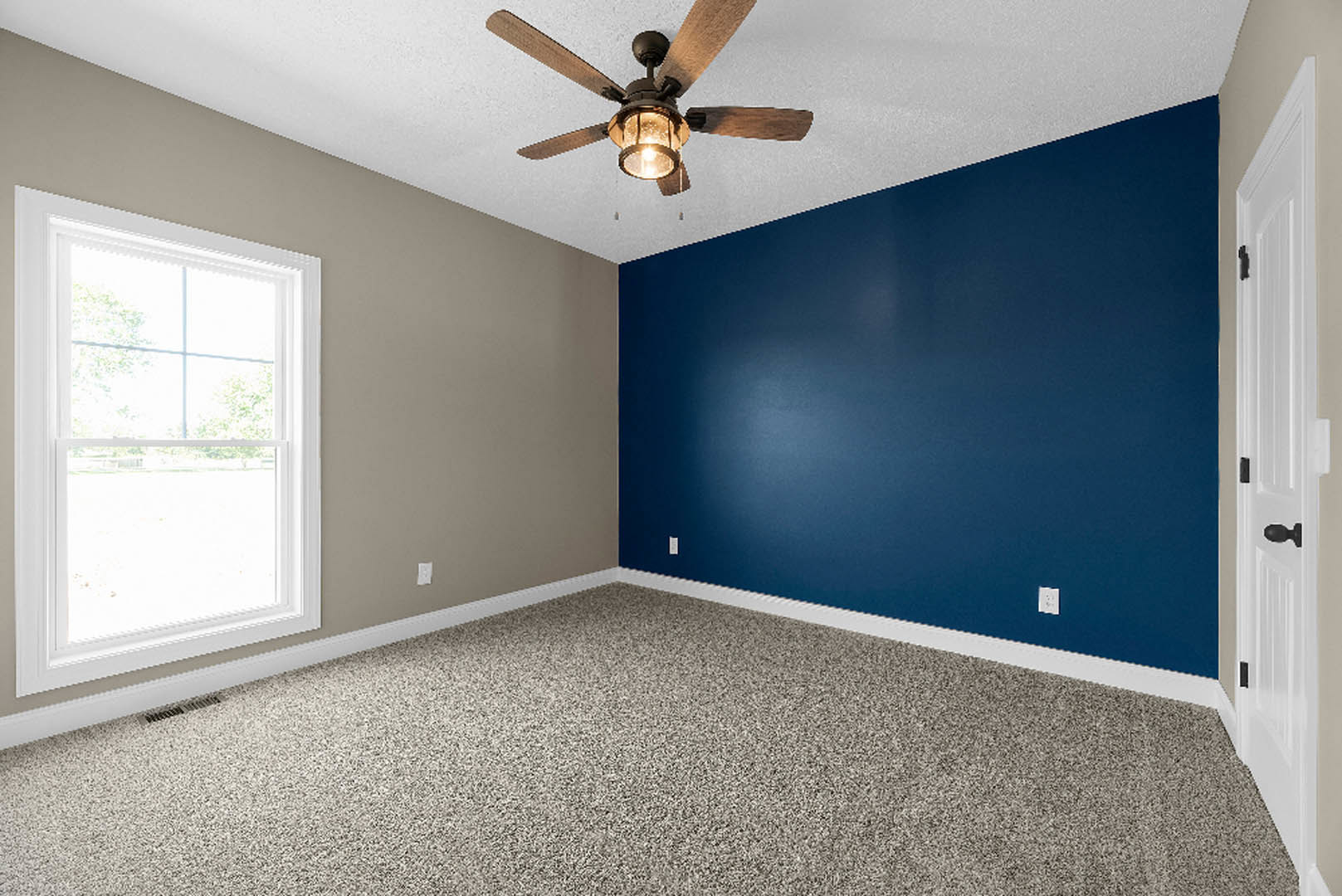 Bedroom with blue painted walls, white trim and molding, ceiling fan with light fixture, carpeted floor, white door with black handle, window overlooking grassy field