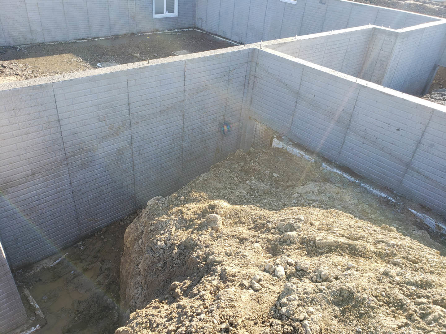 Brick exterior wall with a hole, large mound of soil and rocks beside foundation, ground covered in dirt, close-up window visible