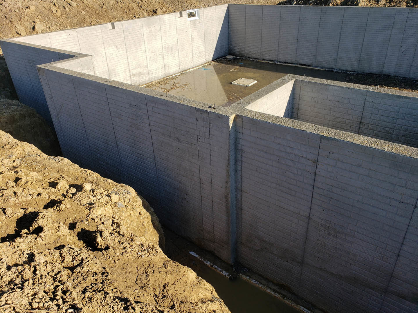 Concrete foundation wall with exposed dirt and a large hole in the ground, outdoor construction site.