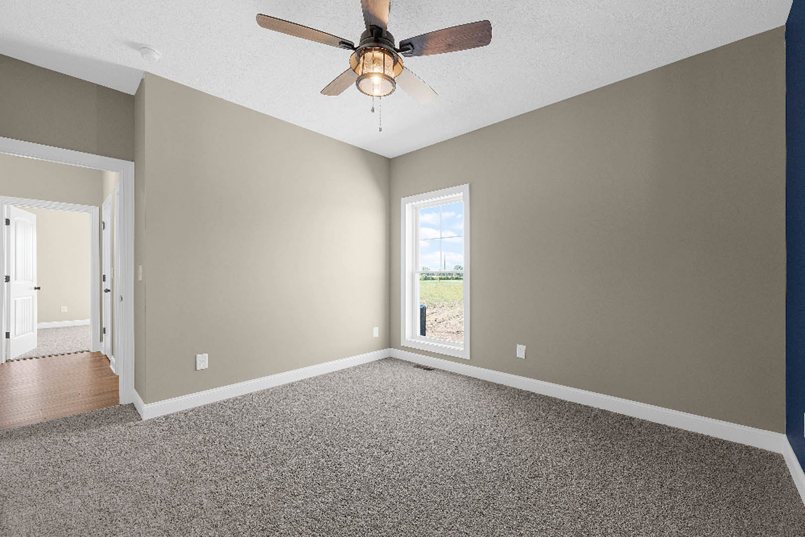 Carpeted room with white walls, ceiling fan with light fixture, large window overlooking grassy field, open white door with black hardware