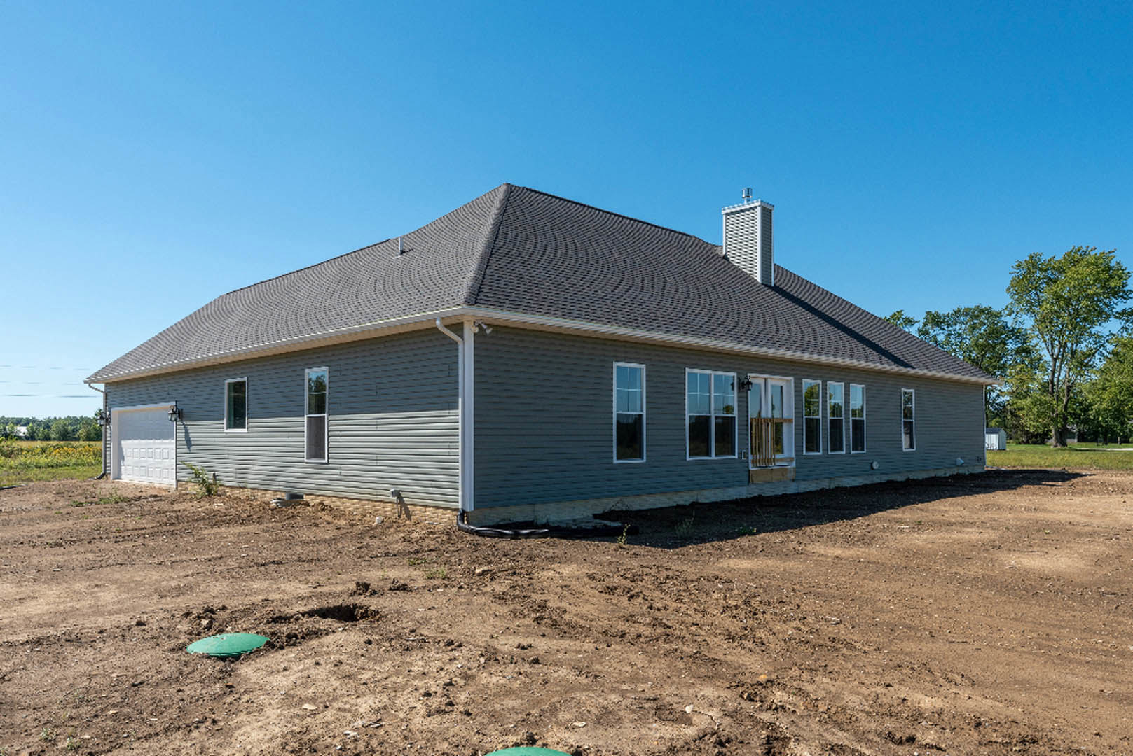 Partially built house with exposed framing, shingled roof, dirt lot in foreground, blue sky above, scattered trees in background, green construction equipment near foundation