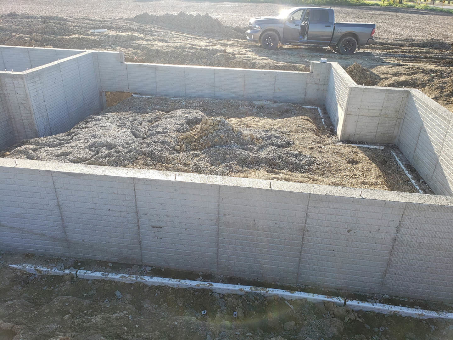 Open truck parked beside concrete foundation, exposed dirt and pipe, construction materials scattered on site, grassy edge visible