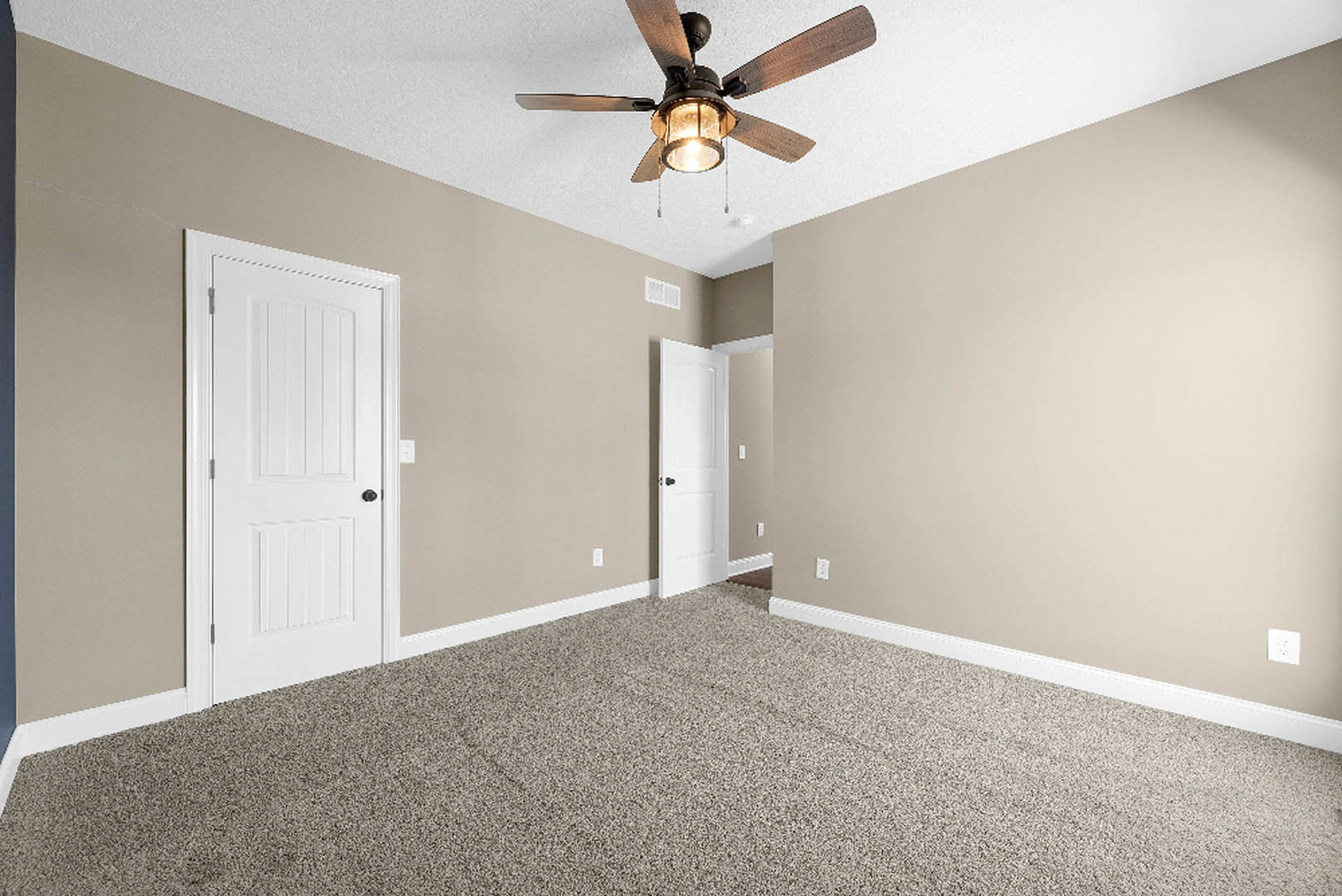 Ceiling fan with light fixture above carpeted floor, white door with black knob, white outlet and vent on light-colored plaster wall, simple molding along baseboard.