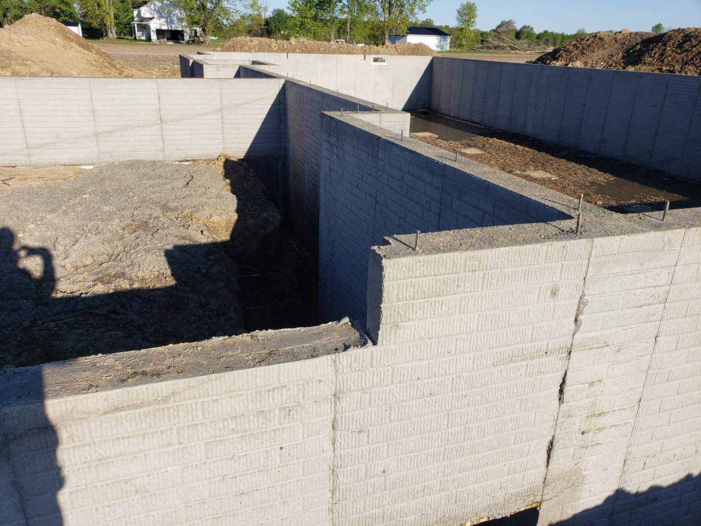 Concrete foundation with a hole in the wall, surrounded by dirt and construction debris, field with trees and parked car in background, white house partially visible, shadow of